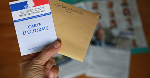 Une main porte une carte électorale accompagnée d'une enveloppe marron.