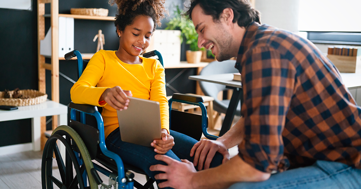 Petite fille noire avec un teeshirt jaune, assise sur un fauteuil roulant, montrant l'écran de sa tablette à un homme blanc portant une chemise à carreau bleus et marrons clairs. Derrière se trouve des étagère en bois sur la gauche et un bureau sur la droite.