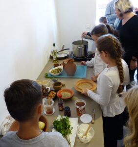 Photo de trois enfants qui préparent les ingrédients pour confectionner un plat. En arrière plan une femme adulte mélange une préparation dans une marmite.