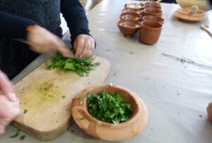 Photo de mains d'adulte découpant des herbes aromatiques sur une planche en bois. Devant se trouve un pilon avec d'autres herbes prêtent à être écrasées