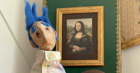 Photo d'une marionnette représentant un homme avec des sourcils et des cheveux bleus, et une blouse d'artiste, devant le célèbre portrait de Mona Lisa, plus célèbrement appelé La Joconde.