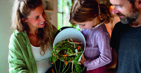 Photo d'une famille dans une cuisine avec une petite fille vidant un sceau d'épluchure de légumes dans un composteur. Elle est accompagnée dans sa démarche de sa mère, à gauche, et de son père, à droite.